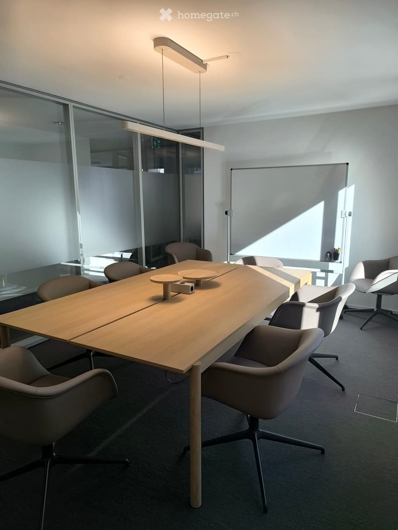 Modern conference room with a long wooden table, several chairs, and a large screen on the wall. The room features large windows and sleek, contemporary lighting fixtures.