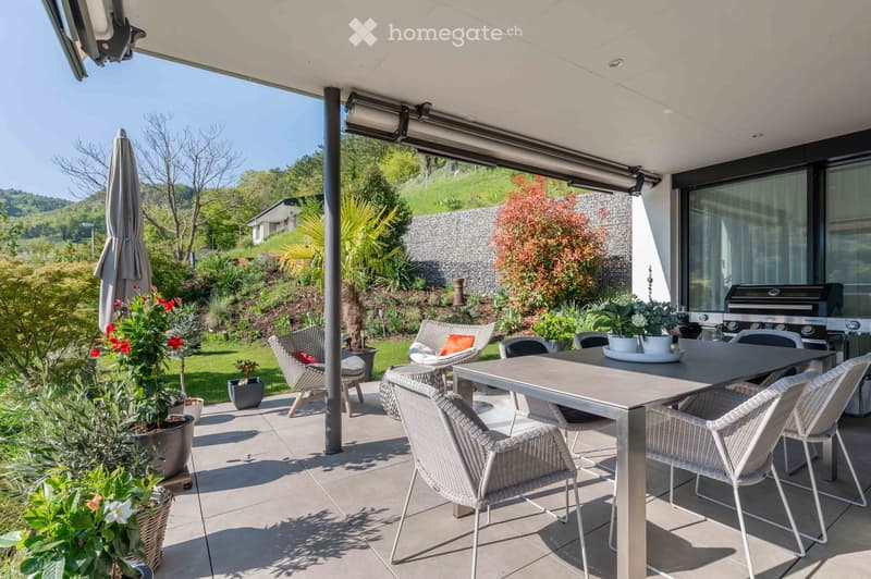 A modern outdoor terrace with a dining table, chairs, umbrella, and potted plants, overlooking a landscaped garden with a barbecue and scenic view.