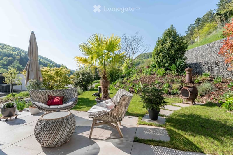 A well-maintained garden with a patio area featuring wicker chairs, a round table, and a large umbrella. The garden includes various plants, a palm tree, and a stone wall in the background.