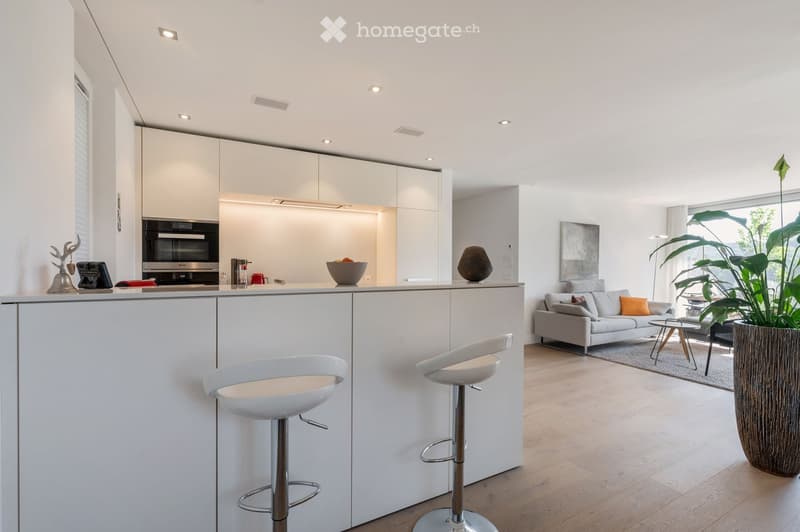 Modern, open-plan living and dining area with white cabinets, a grey sofa, two white bar stools, a coffee table, and a large potted plant near the window.