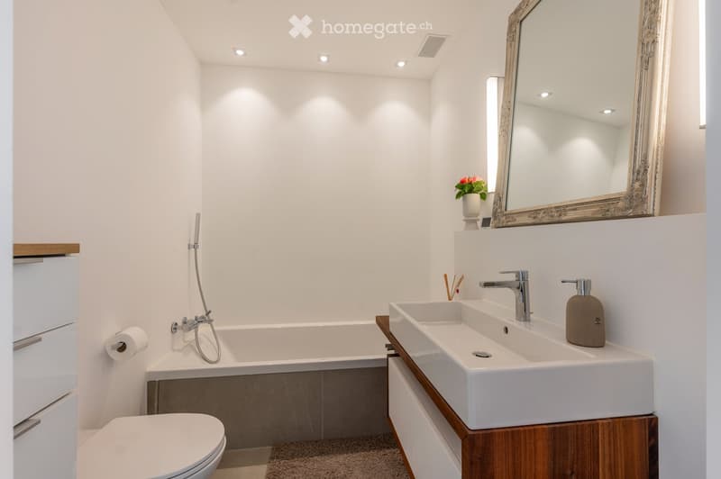 Modern bathroom with a white bathtub, toilet, double sink, and a large ornate mirror. Features include a wooden vanity, a small flower vase, and a soap dispenser.