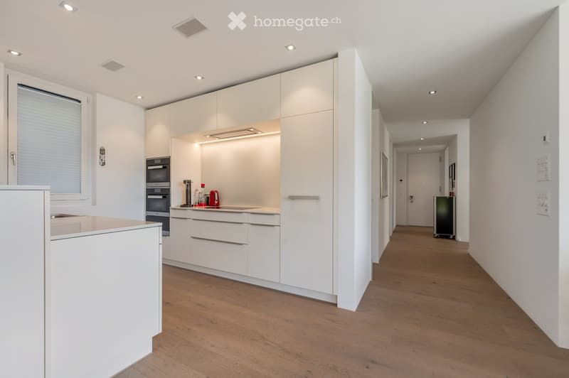 Modern kitchen with white cabinets, wooden flooring, and built-in appliances including an oven and coffee maker. The hallway leads to another room with a visible refrigerator.