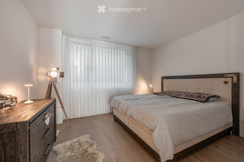 Modern bedroom with a large bed, wooden dresser, lamp, furry rug, and tripod lamp near the window.