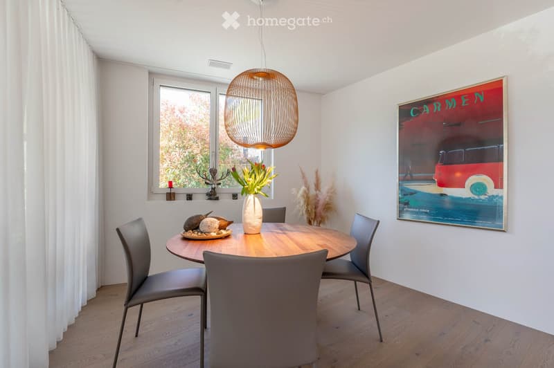 Modern dining area with a round wooden table surrounded by grey chairs, a decorative vase with flowers, and a large poster on the wall. A wicker pendant light hangs from the ceiling, and the room features large windows with white curtains.