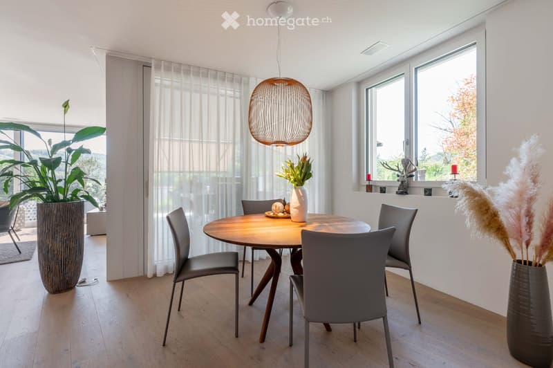 Modern dining room with a round wooden table, six grey chairs, a large pendant light, and decorative vases with plants and candles on the windowsill.