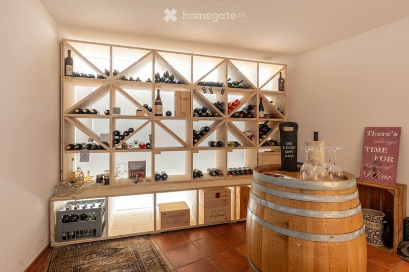 A wine cellar with a geometric wooden wine rack filled with bottles, a wooden barrel as a table with wine glasses, and various wine boxes and jars on display.