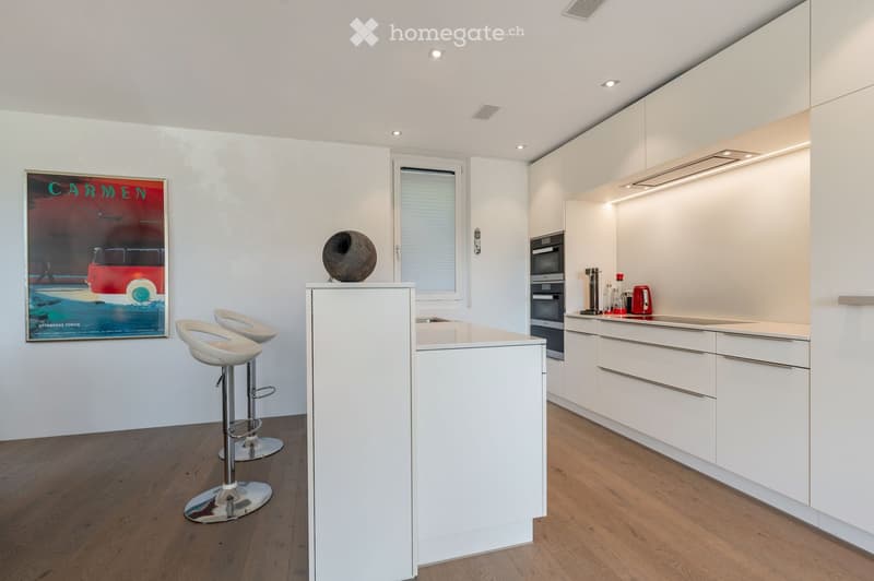 Modern kitchen with white cabinets, a central island, two white bar stools, and a built-in oven. The space features wooden flooring, recessed lighting, and a large window with blinds.