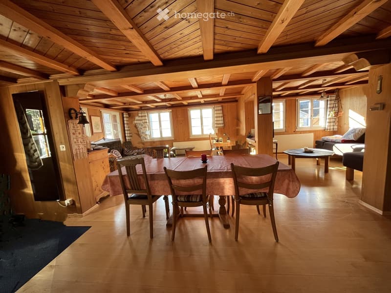 Cozy living and dining area with wooden beams, exposed wooden ceiling, large windows, dining table with chairs, sofa, and television.