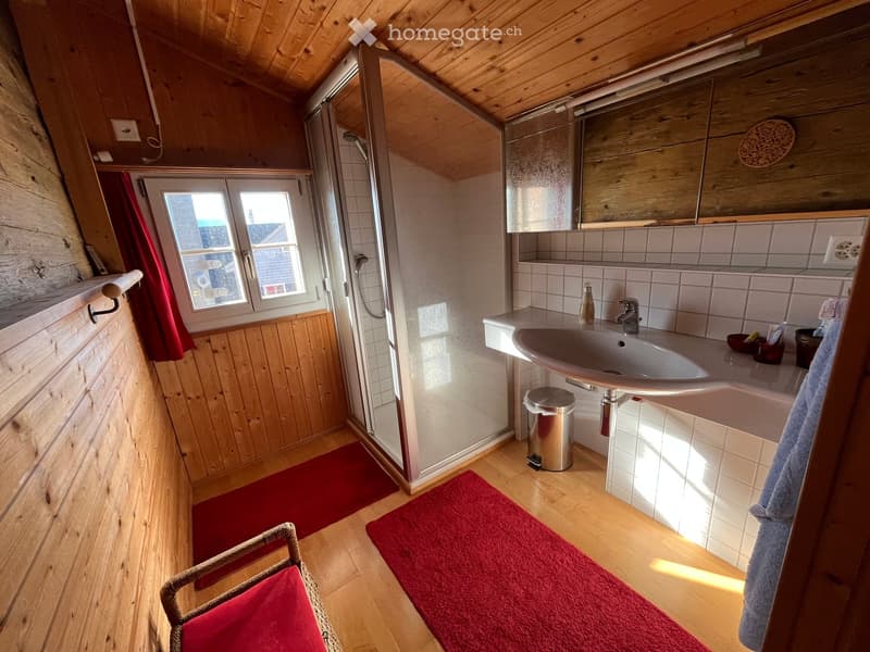 Cozy bathroom with wooden walls and floor, a sink with a mirror, a shower area, and a small red chair near the window.