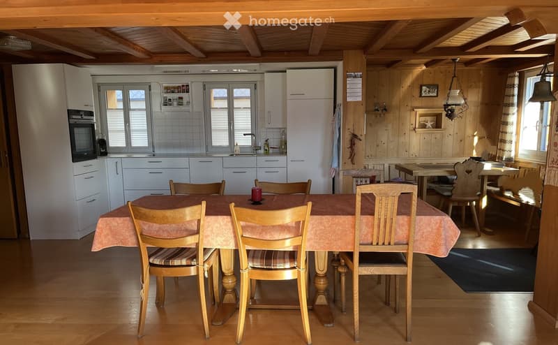Cozy dining area with wooden chairs and a table covered in a pink tablecloth, adjacent to a modern kitchen with white cabinets, a refrigerator, and an oven. Wooden walls and ceiling beams add warmth to the space.