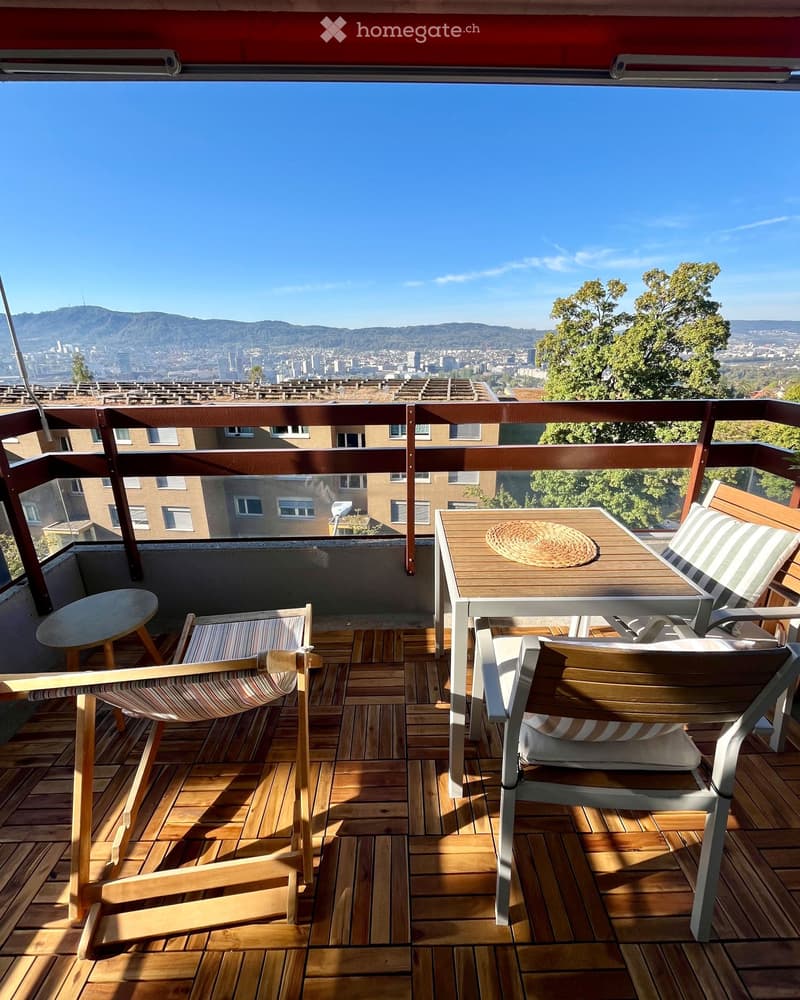 Balcony with wooden decking, table, chairs, and a view of the city and mountains.