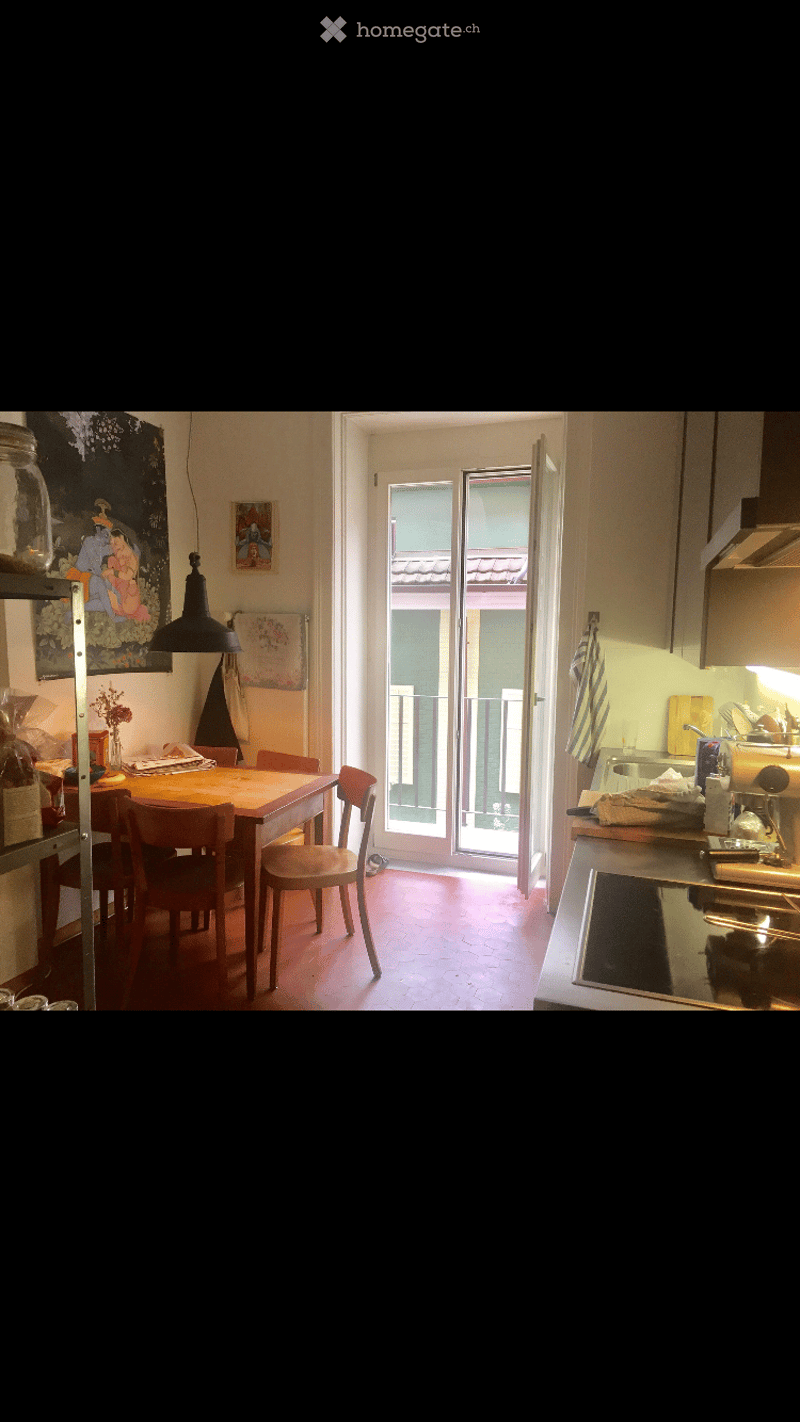 A cozy, well-lit room with a dining area and a small kitchen. The dining area features a wooden table with four chairs, and the kitchen has a counter with a sink, oven, and fridge. A large window with a balcony view is visible, and the room is decorated w