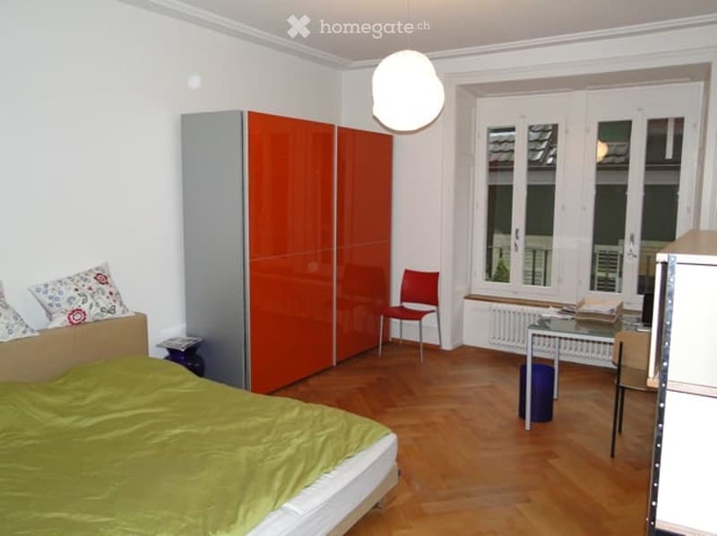 Bright bedroom with wooden flooring, large windows, a bed with green bedding, a red wardrobe, a red chair, and a small desk with a lamp.