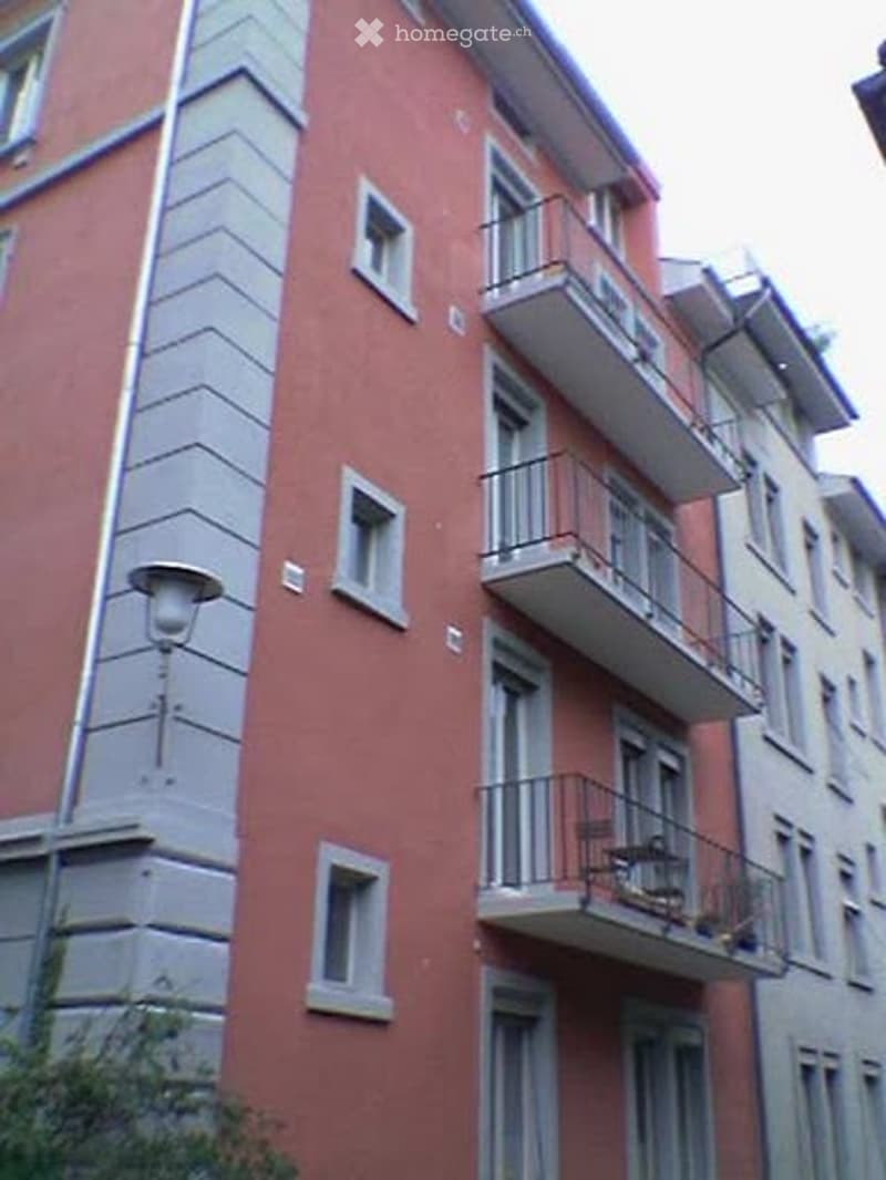 Multi-story residential building with red brick facade, white window frames, and multiple balconies with metal railings.