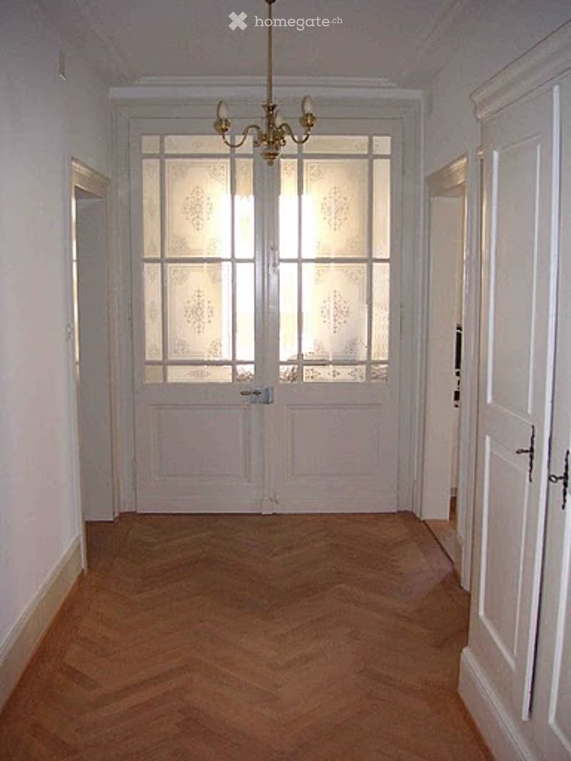 Empty hallway with wooden flooring, white walls, double doors with frosted glass panels, and a chandelier hanging from the ceiling.