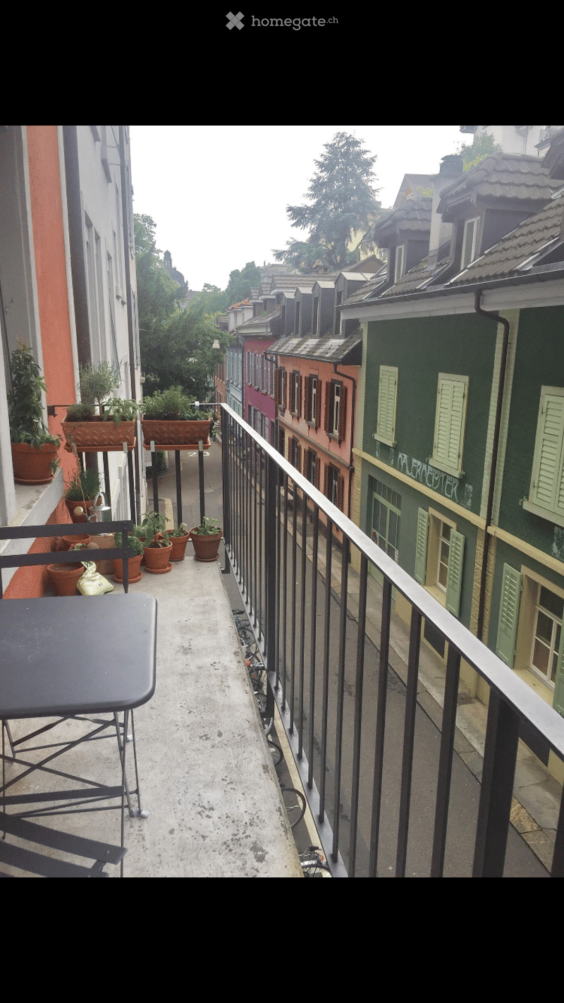 Balcony with a table, chairs, and several potted plants, overlooking a colorful row of houses.