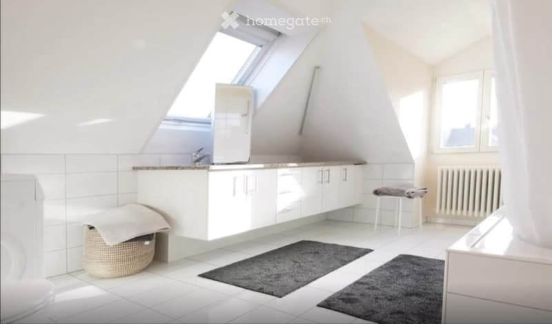 Bright, modern bathroom with white tiled walls and floor, a large skylight window, a sink with a mirror, built-in cabinets, a small stool with towels, a radiator, and a wicker basket on the floor.