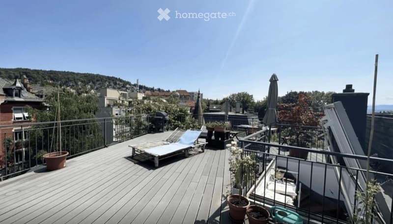Wooden deck with lounge chair, small table, potted plants, and umbrella, overlooking a cityscape with buildings and greenery.