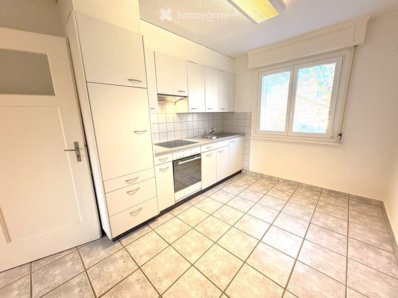 Modern kitchen with white cabinets, tiled floor, sink, oven, and window with view of trees.