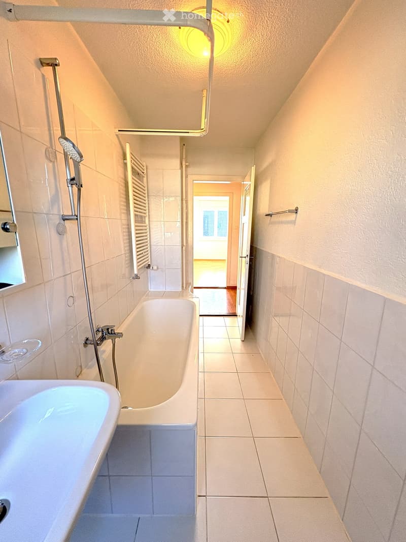 Bathroom with a bathtub, shower, mirror, and towel rack. The space features tiled walls and a slanted ceiling with a circular light fixture.