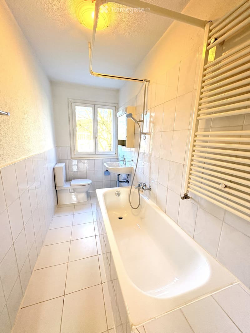 Bright bathroom with white tiled walls and floor, featuring a bathtub, toilet, sink, and a radiator on the right side.