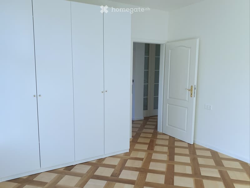 Empty room with white walls, wooden floor, large white wardrobe, and an open door leading to another room.