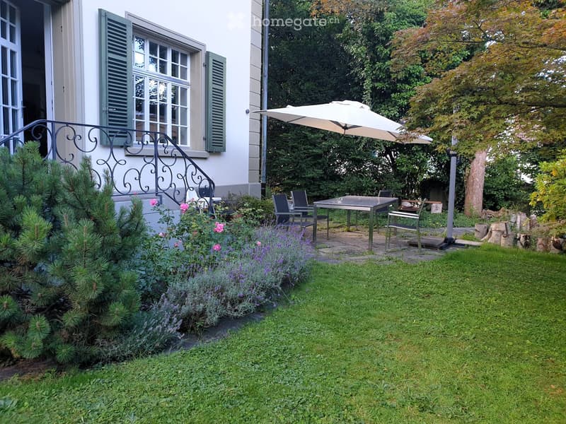 Well-maintained garden with a patio area, outdoor table and chairs, umbrella, and surrounding greenery including shrubs and trees.