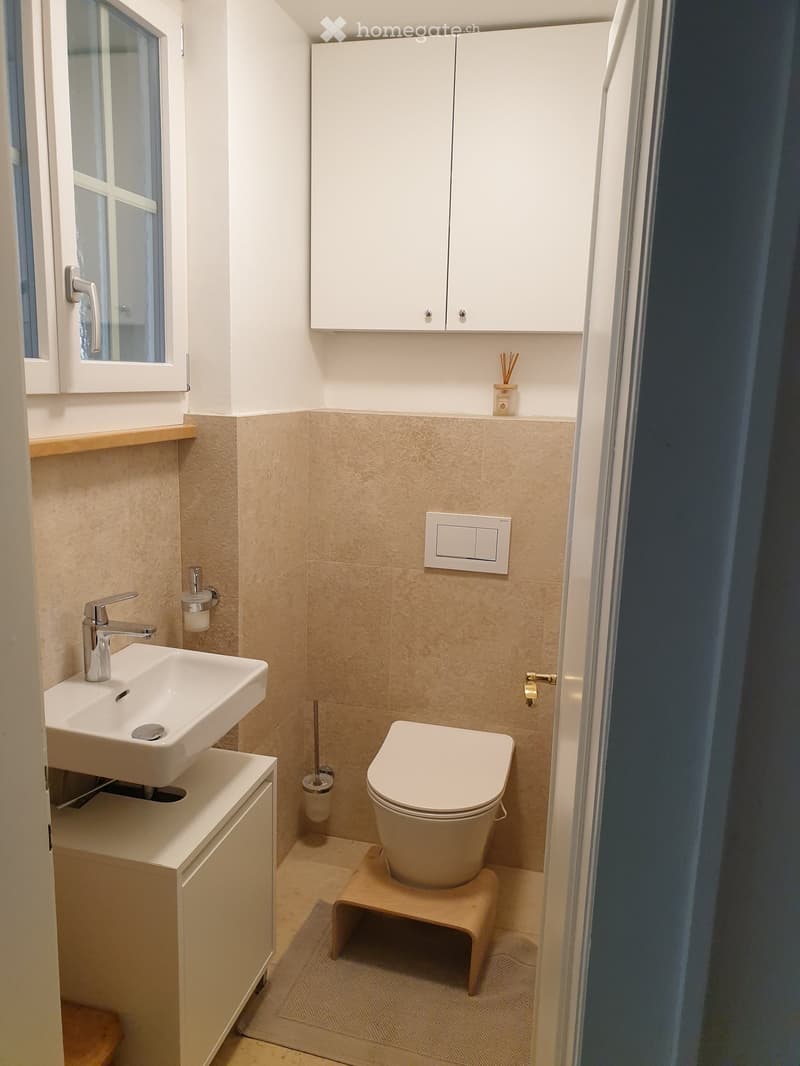 Modern bathroom with a white sink, chrome faucet, toilet, and built-in white cabinets above the sink. Beige tiles and a small wooden step stool are also visible.