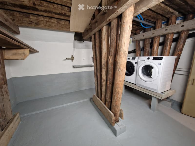 Utility room with exposed wooden beams, concrete floor, and white washing machine and dryer on a wooden platform.