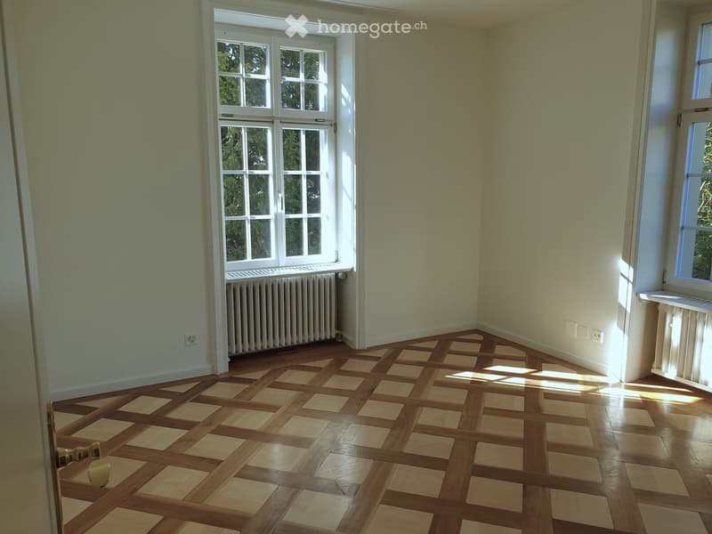 Empty room with light green walls, wooden parquet flooring, and large windows allowing natural light. Radiators are visible under the windows.
