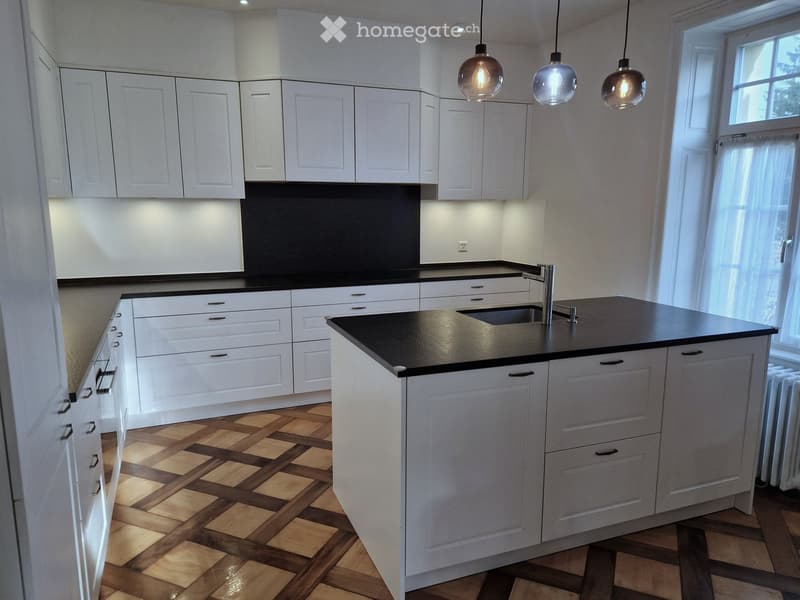 Modern kitchen with white cabinets, black countertops, and a central island. Features pendant lights and a window with curtains.