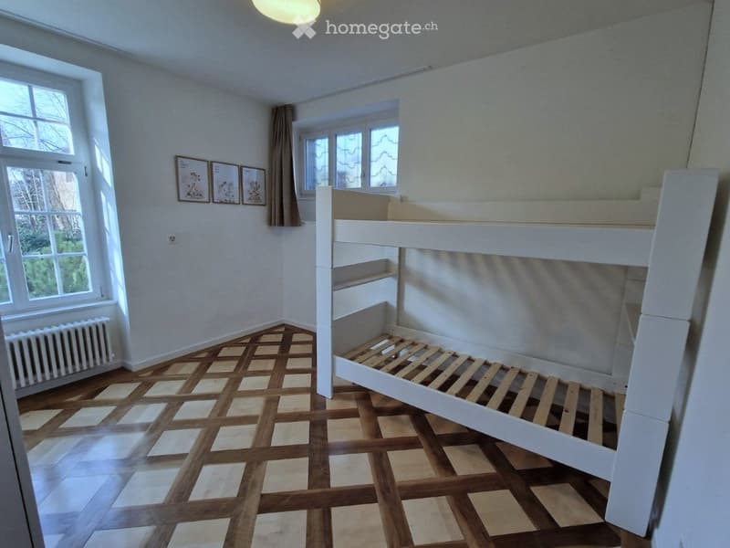 Bright room with wooden floor, large windows, white bunk bed, radiator, and picture frames on the wall.