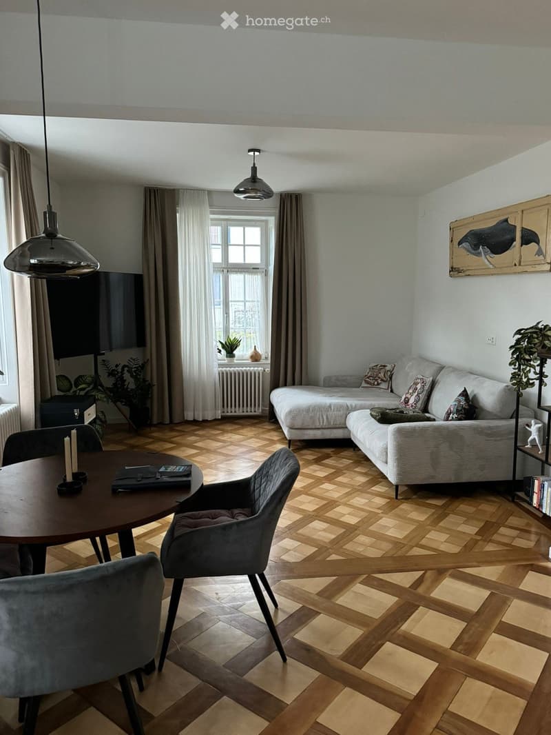 Modern living room with wooden parquet flooring, large window with curtains, grey sofa, armchair, round table, pendant lights, and decorative items including a painting and plants.