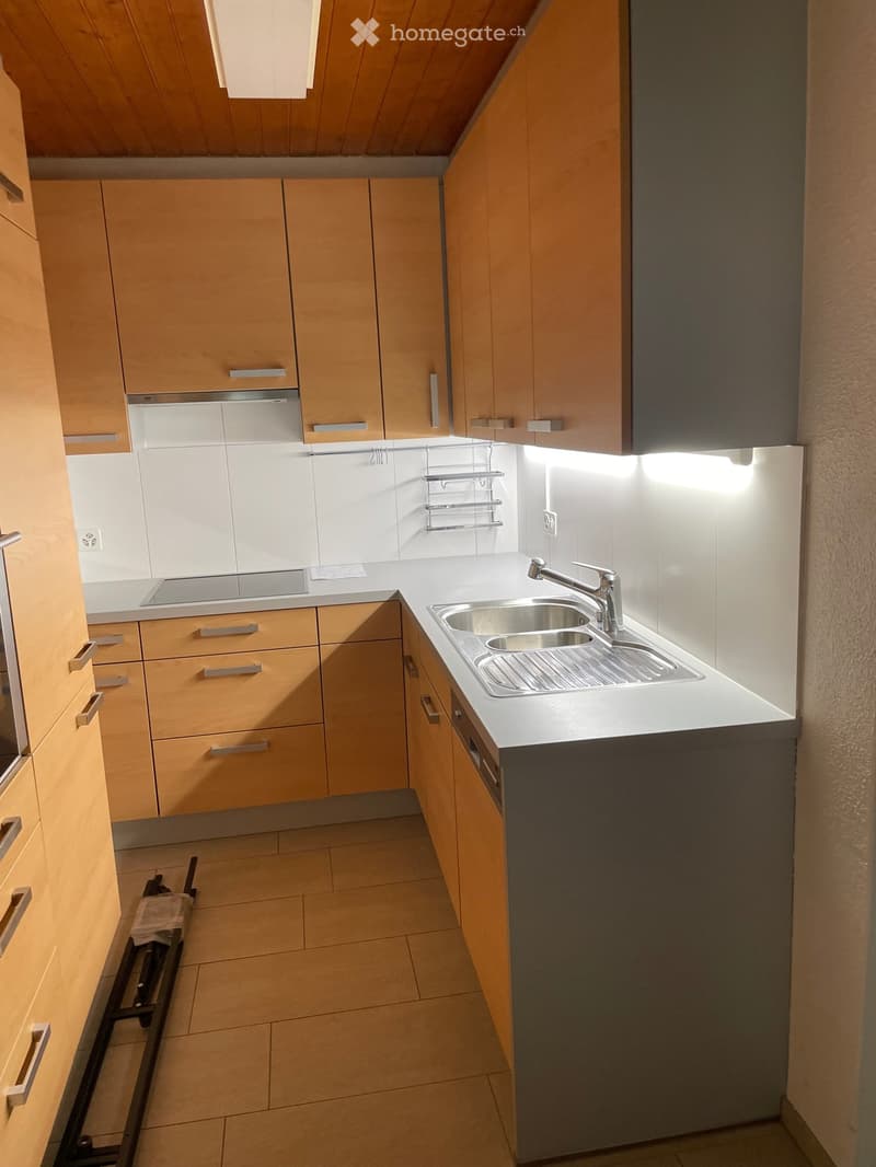 Modern kitchen with light wooden cabinets, stainless steel sink, and built-in oven. The space features a clean, minimalist design with a white countertop and tiled floor.