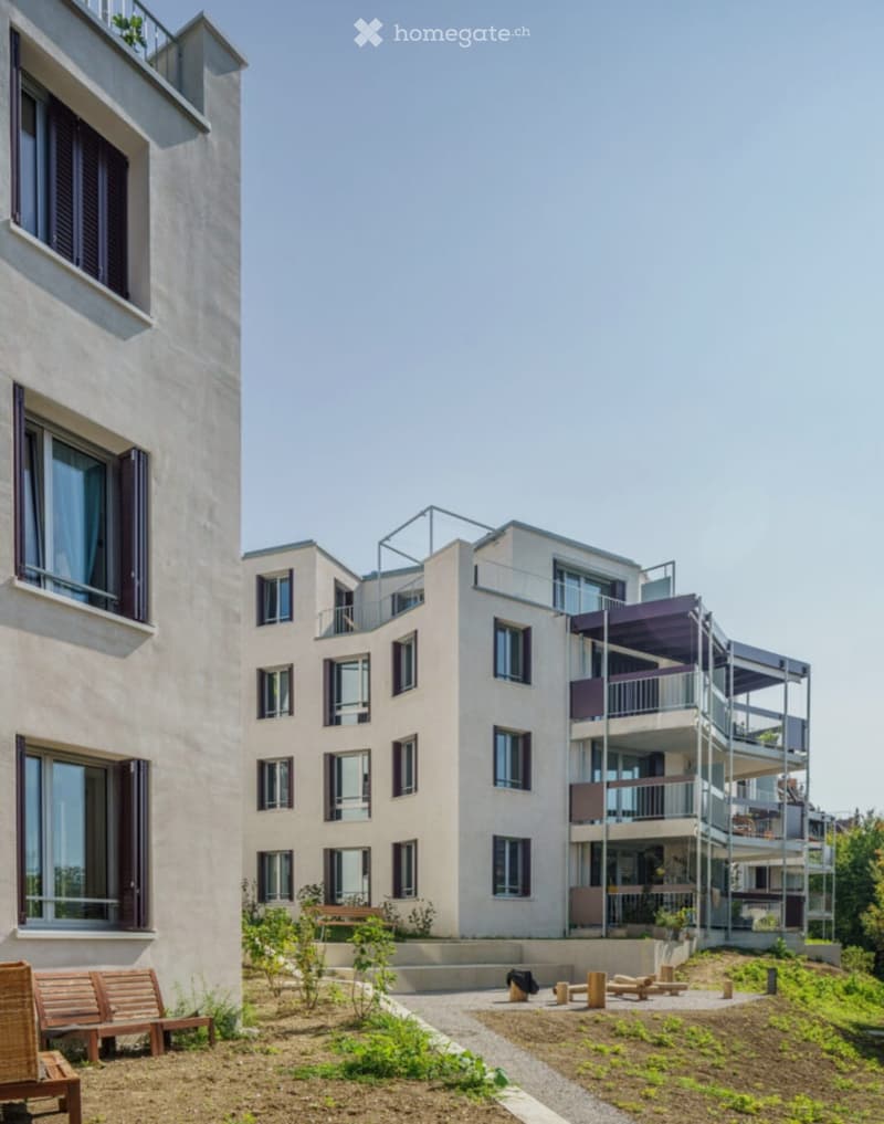 Modern multi-story residential building with balconies and large windows, surrounded by a landscaped garden area with benches and small wooden structures.