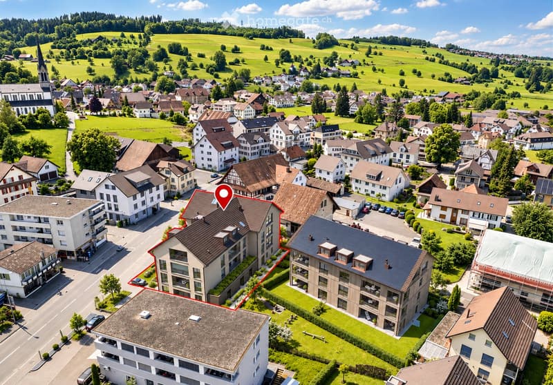 Aerial view of a residential area with multiple houses, green fields, and a church in the background.