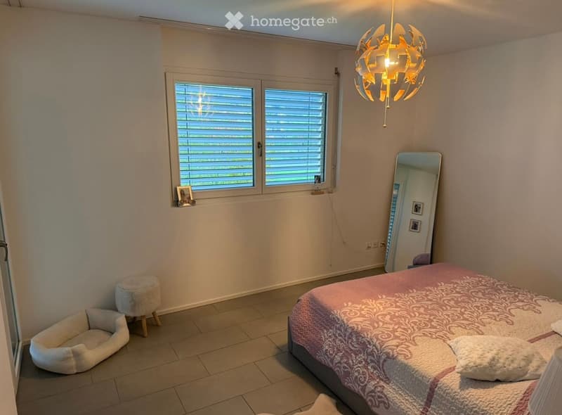 Well-lit bedroom with a double bed, pink and white bedding, a small stool, a large mirror, and a pet bed. The room features large windows with blinds and a modern chandelier.