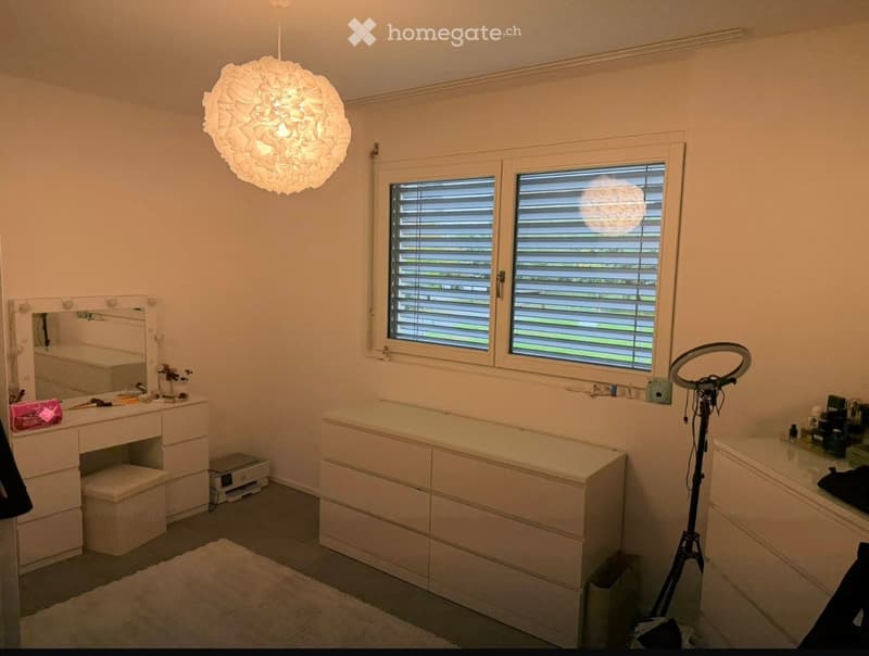 Modern bathroom with white walls, large window with blinds, vanity with mirror, stool, and various toiletries, a large light fixture, and a tripod with a ring light.