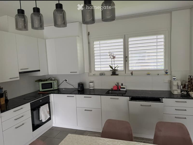 Modern kitchen with white cabinets, black countertops, and stainless steel appliances. Features include a microwave, oven, dishwasher, coffee maker, and a sink with a faucet. There are also chairs and a table in the foreground.