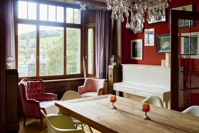 Elegant room with red walls, large windows overlooking a green landscape, a wooden dining table with chairs, a white piano, and decorative candles.