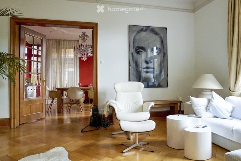 Modern living room with white furniture, including a sofa, coffee table, armchair, and a small table. A large portrait hangs on the wall, and a chandelier is visible in the adjacent dining area.