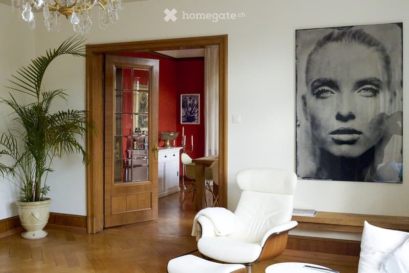 A modern living room with a white leather sofa, a matching chair, a small table, a large picture frame on the wall, a potted plant, and a chandelier hanging from the ceiling. An open doorway leads to another room with red walls and a dining area.