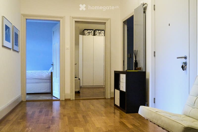 A modern hallway with wooden flooring, a cream-colored sofa, a black cabinet with drawers, and picture frames on the wall. An open doorway leads to a bedroom with a bed visible in the background.