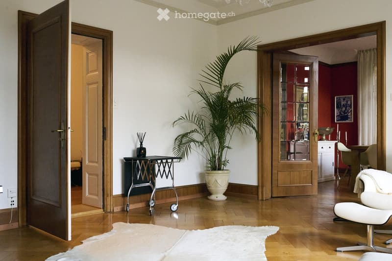 Elegant hallway with wooden flooring, white walls, large potted plant, small table with decorative items, and an open door leading to another room.