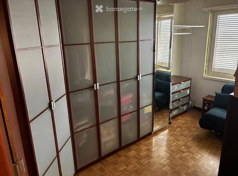 A furnished room with wooden flooring, a large sliding wardrobe, a sofa, a small table, and a drawer unit.