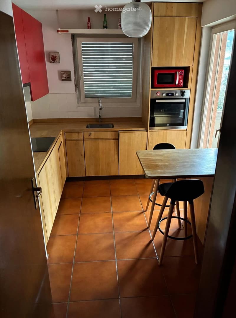 Modern kitchen with wooden cabinets, a red microwave, a built-in oven, a sink with a faucet, a small table with two stools, and a window with blinds.