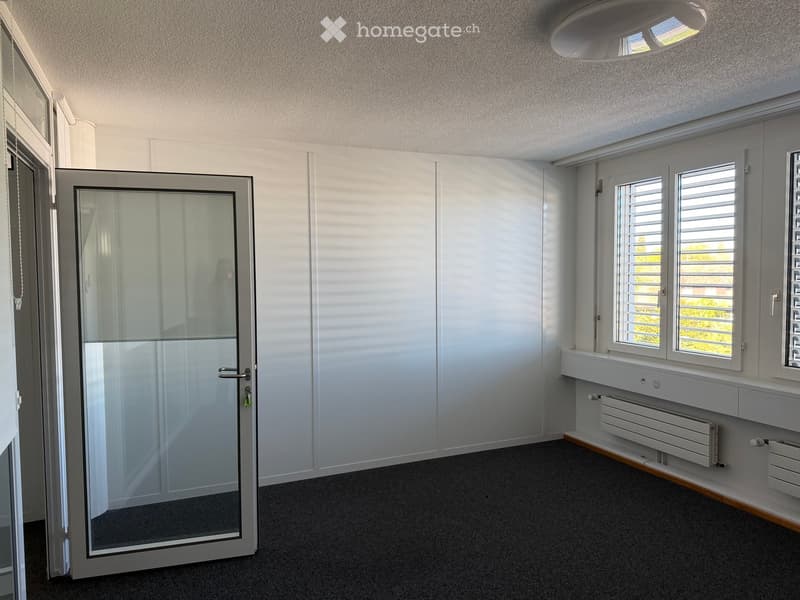 Empty room with white walls, grey carpeted floor, large windows with blinds, and a glass door leading outside.