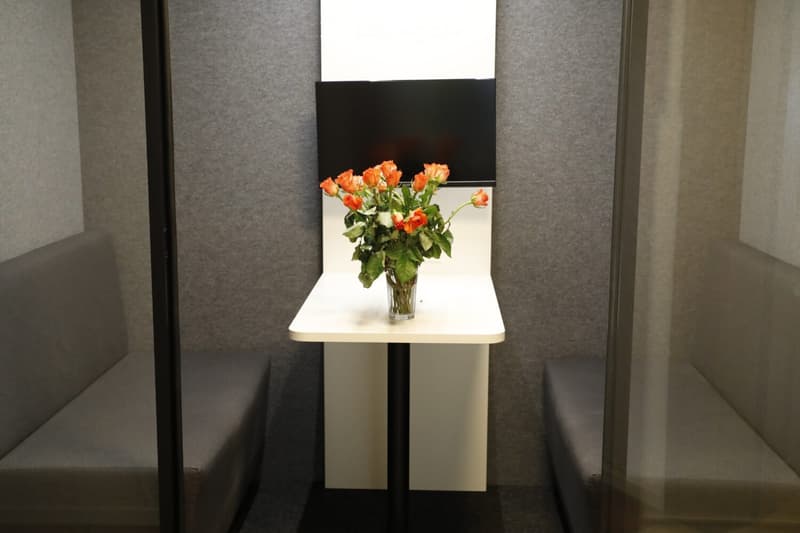 A small, modern room with a white table holding a vase of orange and white flowers, a flat-screen TV mounted on the wall, and grey carpeted flooring.