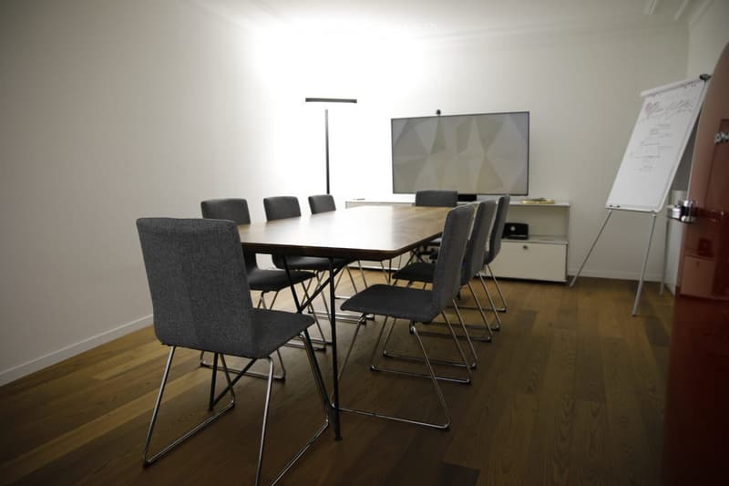 A modern meeting room with a large wooden table, multiple chairs, a screen, and a whiteboard, set on a polished wooden floor.