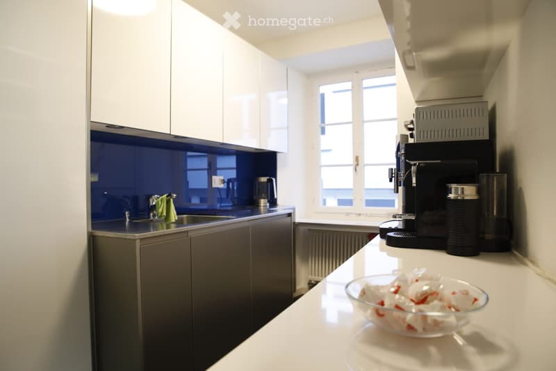 Modern kitchen with white cabinets, dark countertops, and a coffee maker on the counter. A bowl with items is placed on the counter, and a green towel hangs from the faucet.